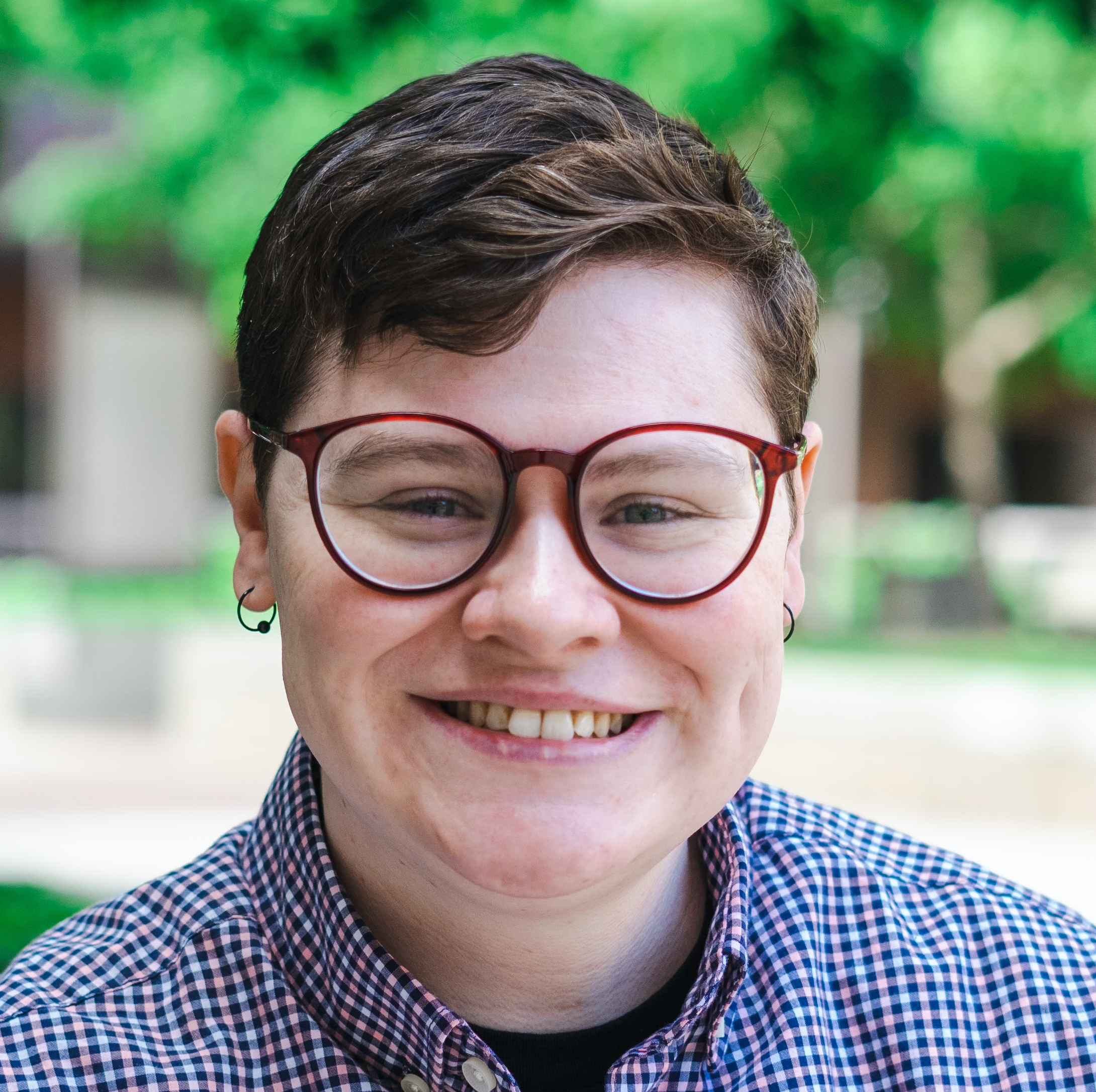 Chest-up image of Ai, a white person with brown hair, blue eyes, and red-framed glasses, who is smiling widely. Zie is wearing a blue and pink plaid shirt.